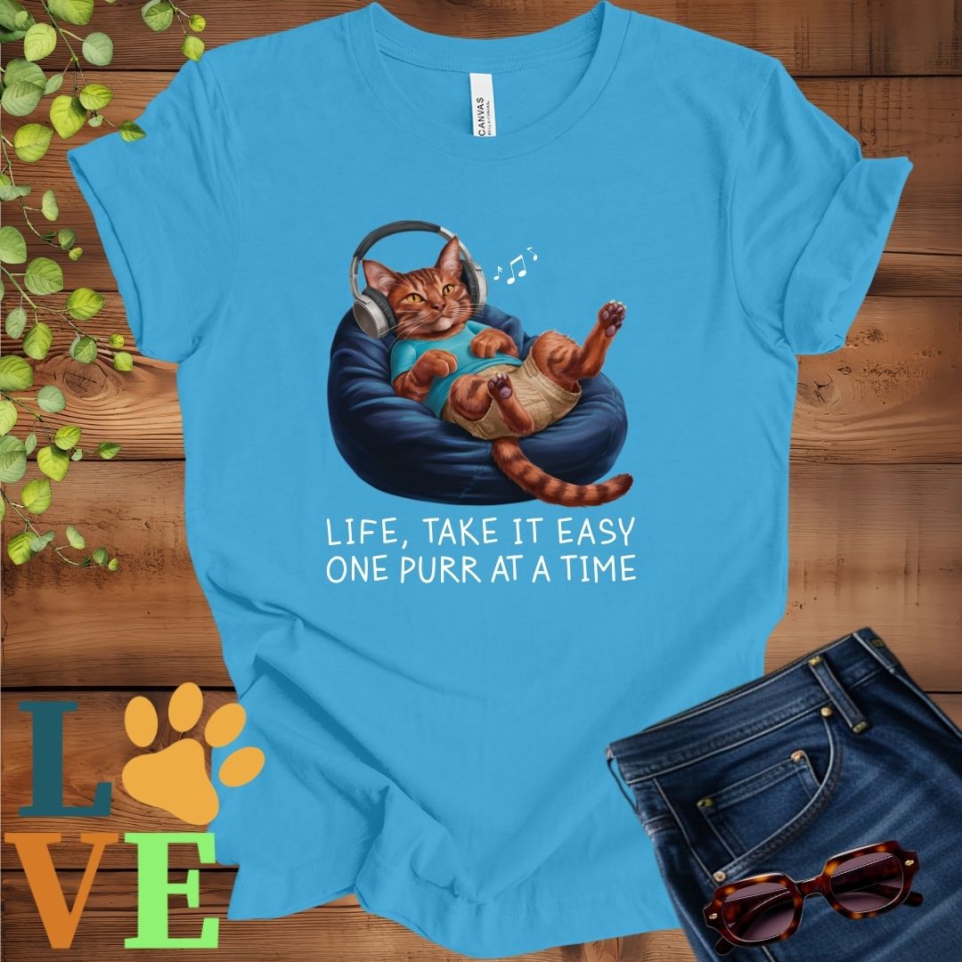 Bright aqua blue t-shirt featuring an anthropomorphic Abyssinian cat relaxing on a bean bag while listening to music through headphones, with the slogan "Life, take it easy. One purr at a time."