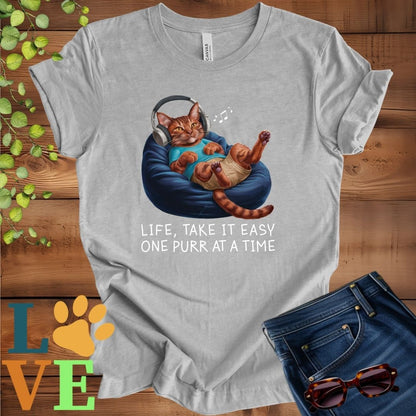 Light grey heather (athletic heather) t-shirt featuring an anthropomorphic Abyssinian cat relaxing on a bean bag while listening to music through headphones, with the slogan "Life, take it easy. One purr at a time."
