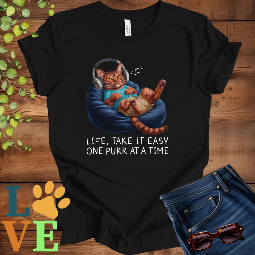 Black t-shirt featuring an anthropomorphic Abyssinian cat relaxing on a bean bag while listening to music through headphones, with the slogan "Life, take it easy. One purr at a time."