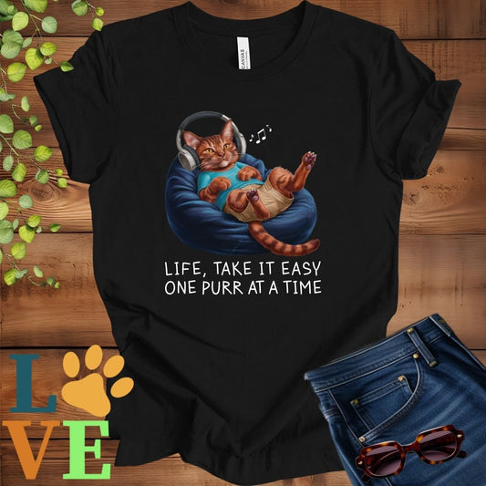 Black t-shirt featuring an anthropomorphic Abyssinian cat relaxing on a bean bag while listening to music through headphones, with the slogan "Life, take it easy. One purr at a time."