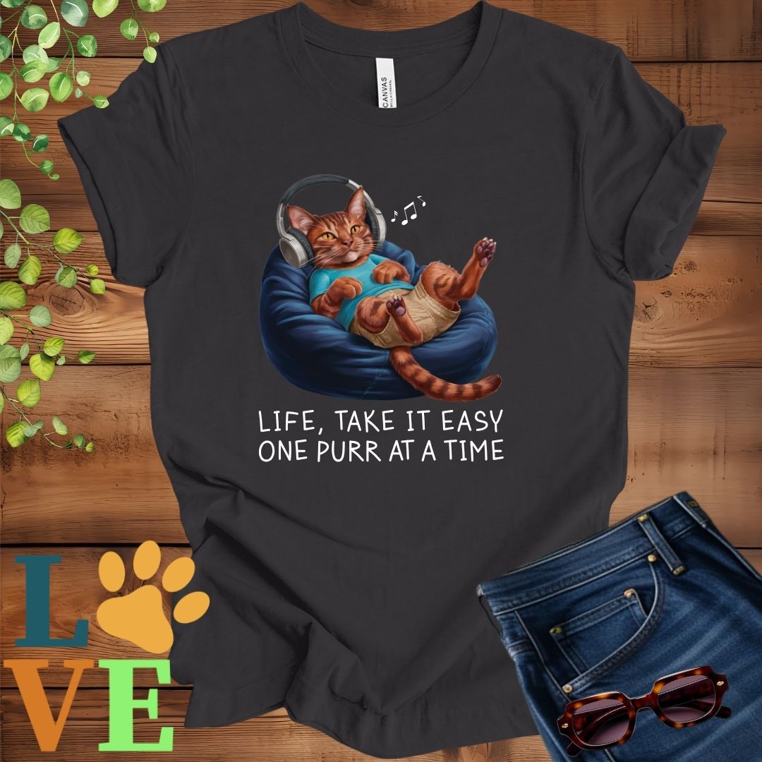 Dark grey heather t-shirt featuring an anthropomorphic Abyssinian cat relaxing on a bean bag while listening to music through headphones, with the slogan "Life, take it easy. One purr at a time."