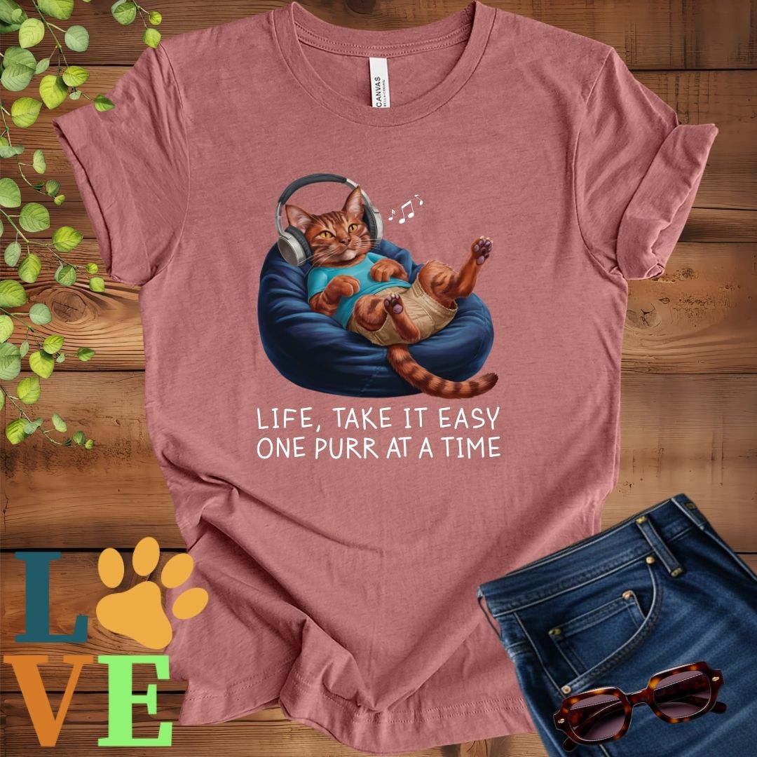 Muted mauve-purple (heather mauve) t-shirt featuring an anthropomorphic Abyssinian cat relaxing on a bean bag while listening to music through headphones, with the slogan "Life, take it easy. One purr at a time."