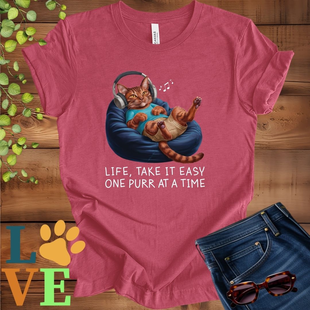 Muted raspberry pink (heather raspberry) t-shirt featuring an anthropomorphic Abyssinian cat relaxing on a bean bag while listening to music through headphones, with the slogan "Life, take it easy. One purr at a time."
