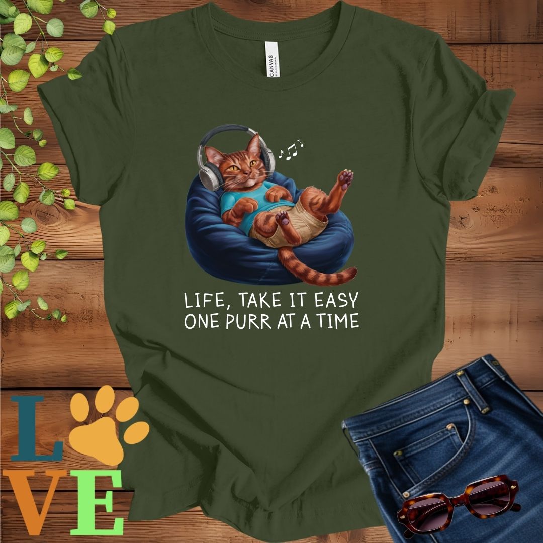 Olive green (military green) t-shirt featuring an anthropomorphic Abyssinian cat relaxing on a bean bag while listening to music through headphones, with the slogan "Life, take it easy. One purr at a time."