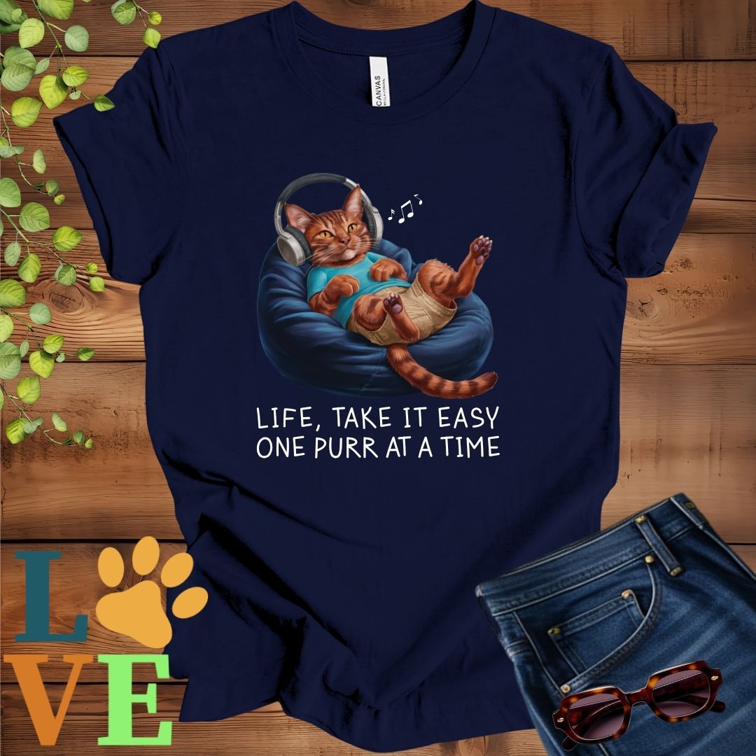 Navy blue t-shirt featuring an anthropomorphic Abyssinian cat relaxing on a bean bag while listening to music through headphones, with the slogan "Life, take it easy. One purr at a time."