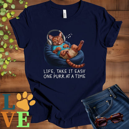 Navy blue t-shirt featuring an anthropomorphic Abyssinian cat relaxing on a bean bag while listening to music through headphones, with the slogan "Life, take it easy. One purr at a time."
