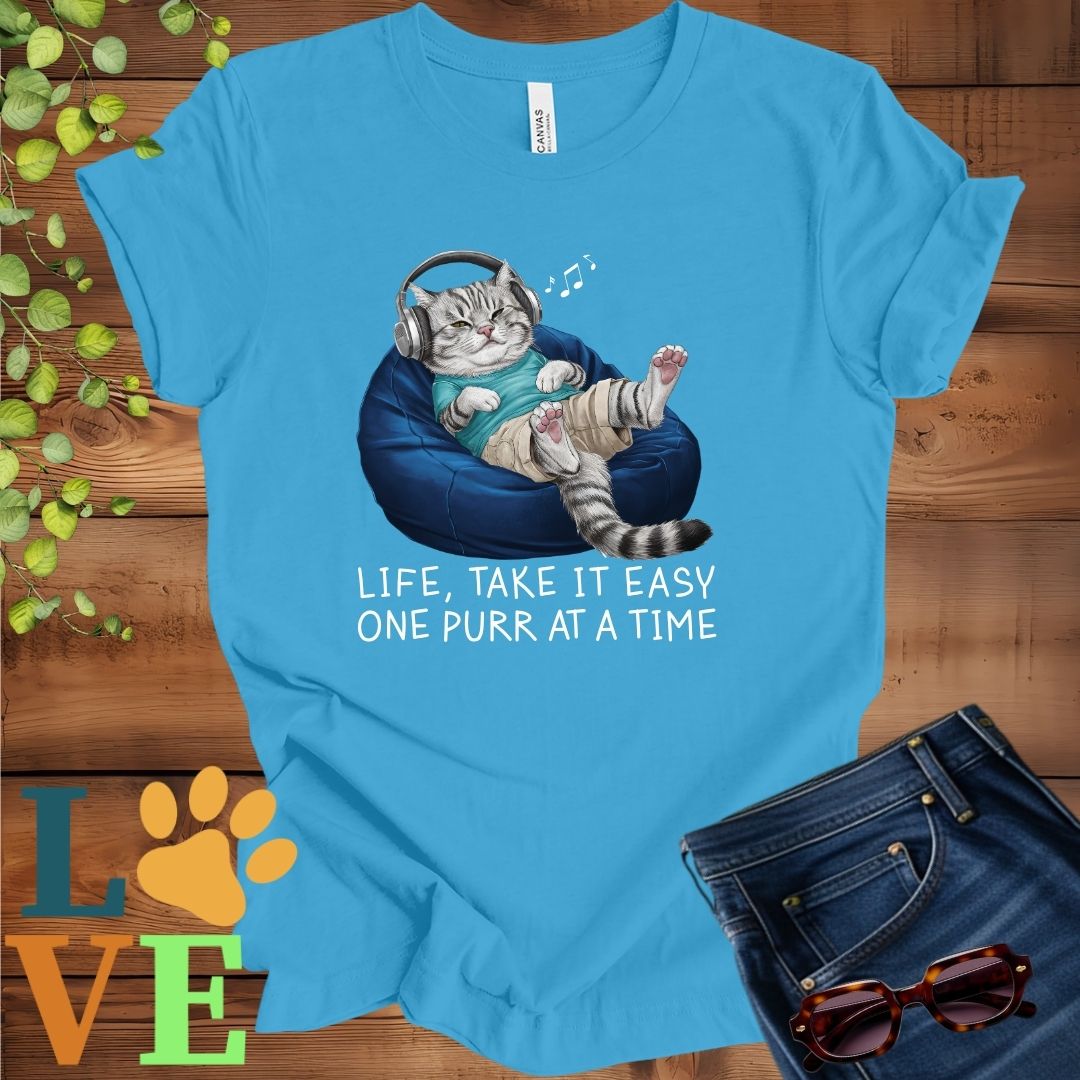 Bright aqua blue t-shirt featuring an anthropomorphic American Shorthair cat relaxing on a bean bag while listening to music through headphones, with the slogan "Life, take it easy. One purr at a time."
