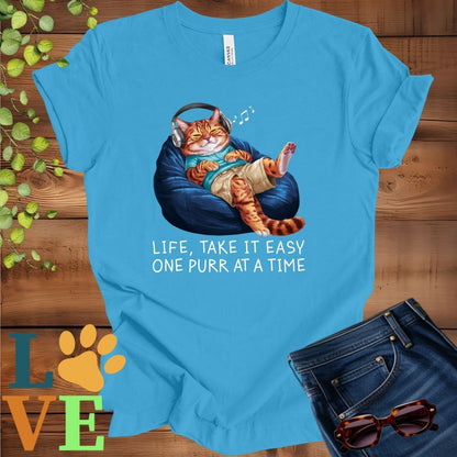 Bright aqua blue t-shirt featuring an anthropomorphic Bengal cat relaxing on a bean bag while listening to music through headphones, with the slogan "Life, take it easy. One purr at a time."
