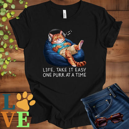 Black t-shirt featuring an anthropomorphic Bengal cat relaxing on a bean bag while listening to music through headphones, with the slogan "Life, take it easy. One purr at a time."