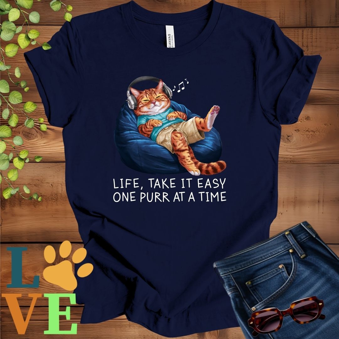 Navy blue t-shirt featuring an anthropomorphic Bengal cat relaxing on a bean bag while listening to music through headphones, with the slogan "Life, take it easy. One purr at a time."