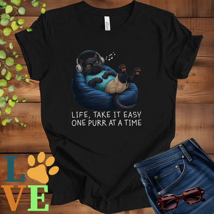 Black t-shirt featuring an anthropomorphic black cat relaxing on a bean bag while listening to music through headphones, with the slogan "Life, take it easy. One purr at a time."