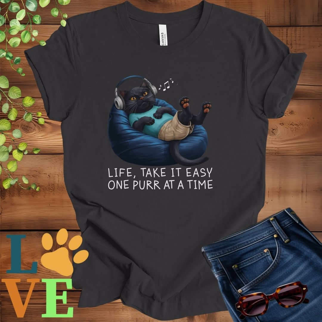 Dark grey heather t-shirt featuring an anthropomorphic black cat relaxing on a bean bag while listening to music through headphones, with the slogan "Life, take it easy. One purr at a time."
