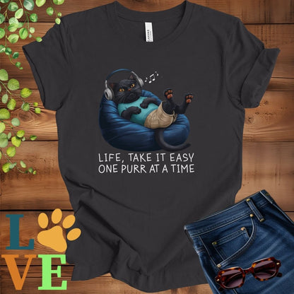 Dark grey heather t-shirt featuring an anthropomorphic black cat relaxing on a bean bag while listening to music through headphones, with the slogan "Life, take it easy. One purr at a time."
