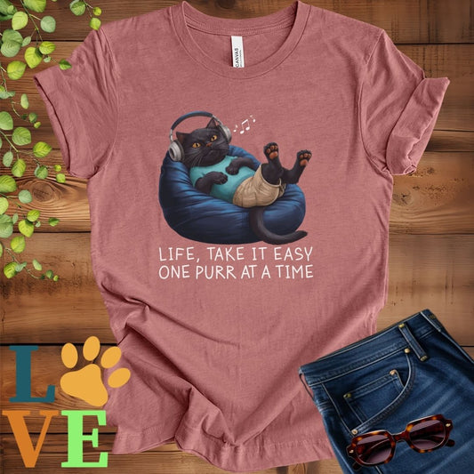 Muted mauve-purple (heather mauve) t-shirt featuring an anthropomorphic black cat relaxing on a bean bag while listening to music through headphones, with the slogan "Life, take it easy. One purr at a time."