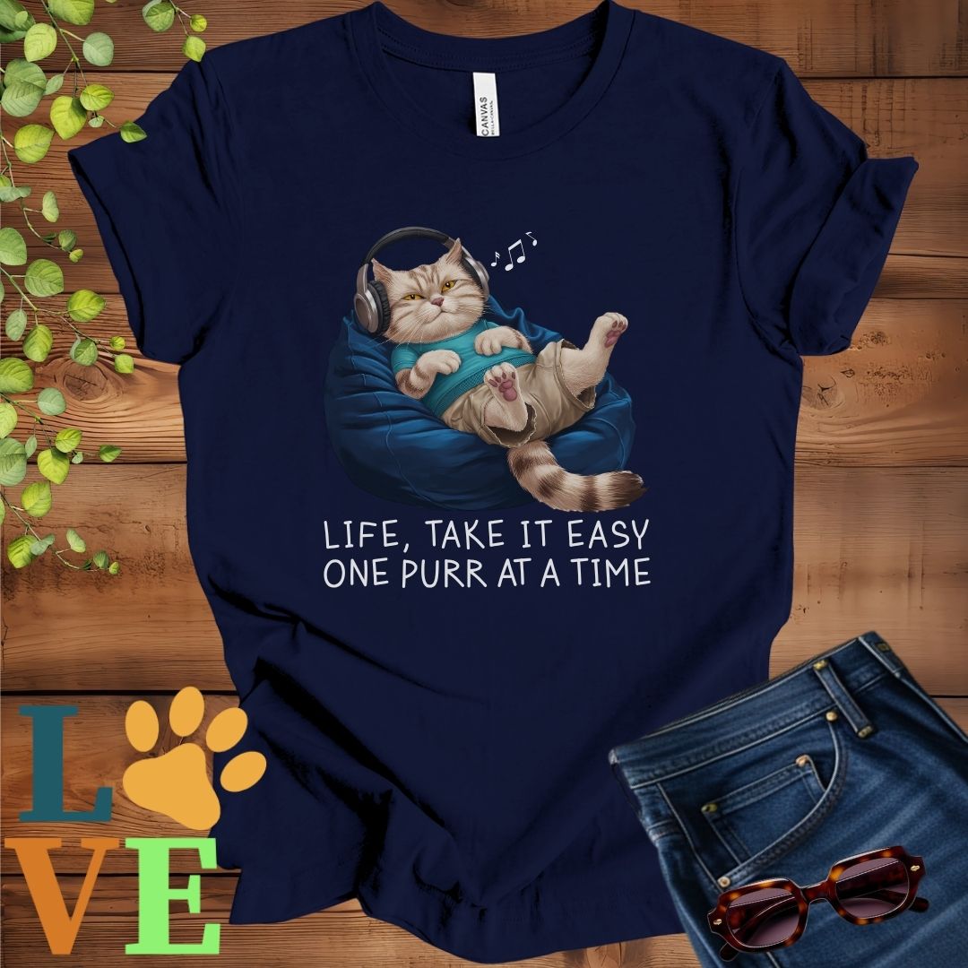 Navy blue t-shirt featuring an anthropomorphic British Shorthair cat relaxing on a bean bag while listening to music through headphones, with the slogan "Life, take it easy. One purr at a time."