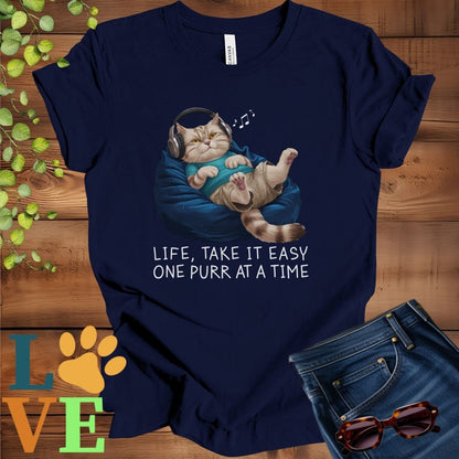 Navy blue t-shirt featuring an anthropomorphic British Shorthair cat relaxing on a bean bag while listening to music through headphones, with the slogan "Life, take it easy. One purr at a time."