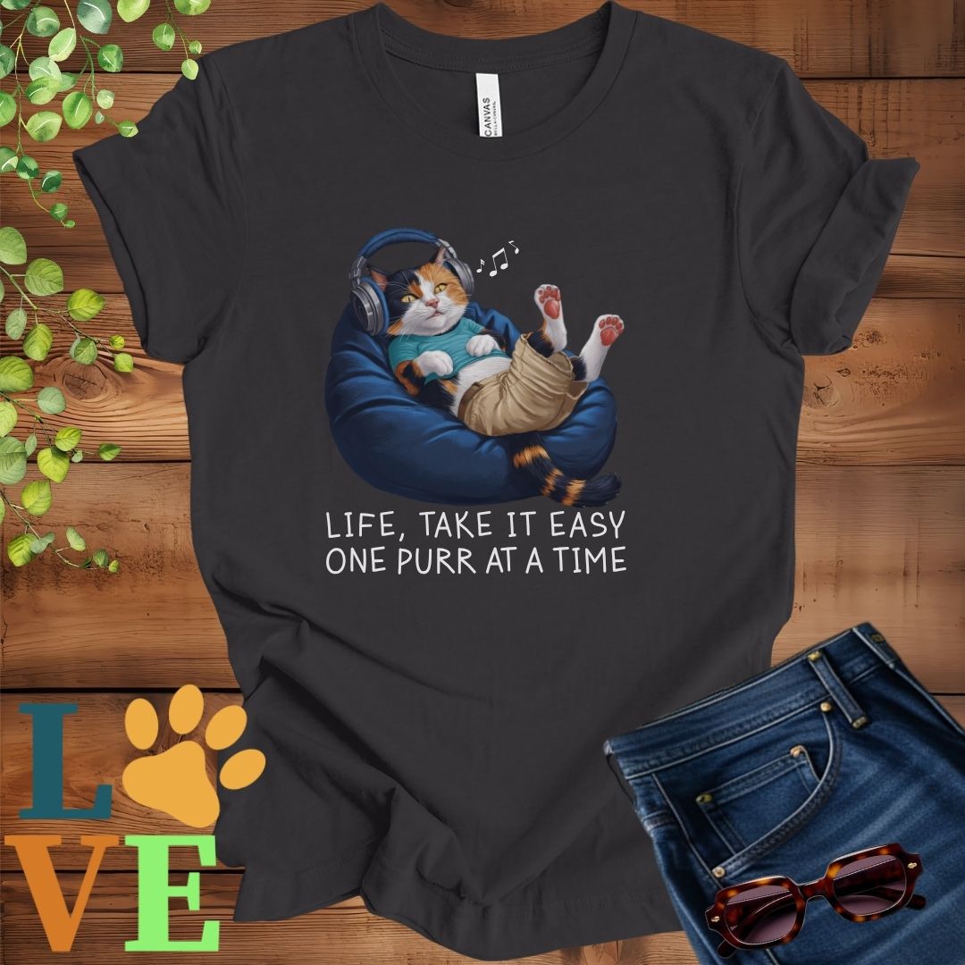 Dark grey heather t-shirt featuring an anthropomorphic calico cat relaxing on a bean bag while listening to music through headphones, with the slogan "Life, take it easy. One purr at a time."