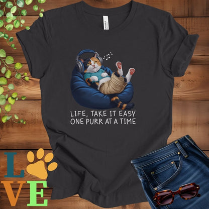 Dark grey heather t-shirt featuring an anthropomorphic calico cat relaxing on a bean bag while listening to music through headphones, with the slogan "Life, take it easy. One purr at a time."