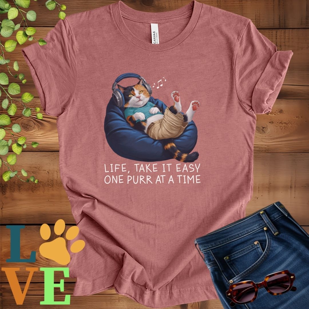 Muted mauve-purple (heather mauve) t-shirt featuring an anthropomorphic calico cat relaxing on a bean bag while listening to music through headphones, with the slogan "Life, take it easy. One purr at a time."