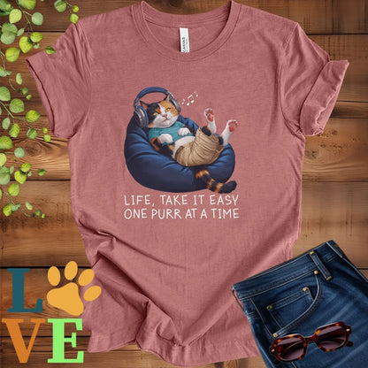 Muted mauve-purple (heather mauve) t-shirt featuring an anthropomorphic calico cat relaxing on a bean bag while listening to music through headphones, with the slogan "Life, take it easy. One purr at a time."