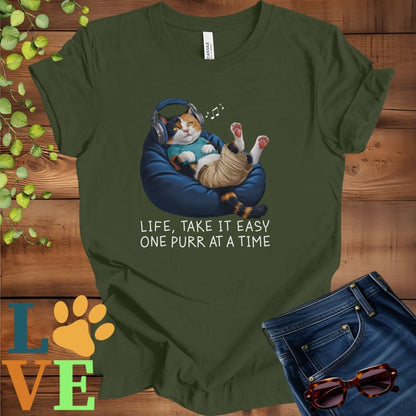 Olive green (military green) t-shirt featuring an anthropomorphic calico cat relaxing on a bean bag while listening to music through headphones, with the slogan "Life, take it easy. One purr at a time."