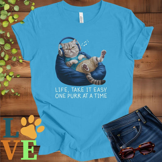 Bright aqua blue t-shirt featuring an anthropomorphic Exotic Shorthair cat relaxing on a bean bag while listening to music through headphones, with the slogan "Life, take it easy. One purr at a time."
