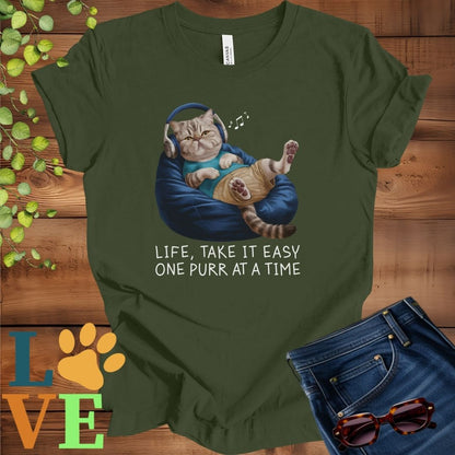 Olive green (military green) t-shirt featuring an anthropomorphic Exotic Shorthair cat relaxing on a bean bag while listening to music through headphones, with the slogan "Life, take it easy. One purr at a time."