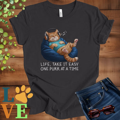 Dark grey heather t-shirt featuring an anthropomorphic ginger cat relaxing on a bean bag while listening to music through headphones, with the slogan "Life, take it easy. One purr at a time."

