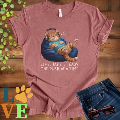 Muted mauve-purple (heather mauve) t-shirt featuring an anthropomorphic ginger cat relaxing on a bean bag while listening to music through headphones, with the slogan "Life, take it easy. One purr at a time."
