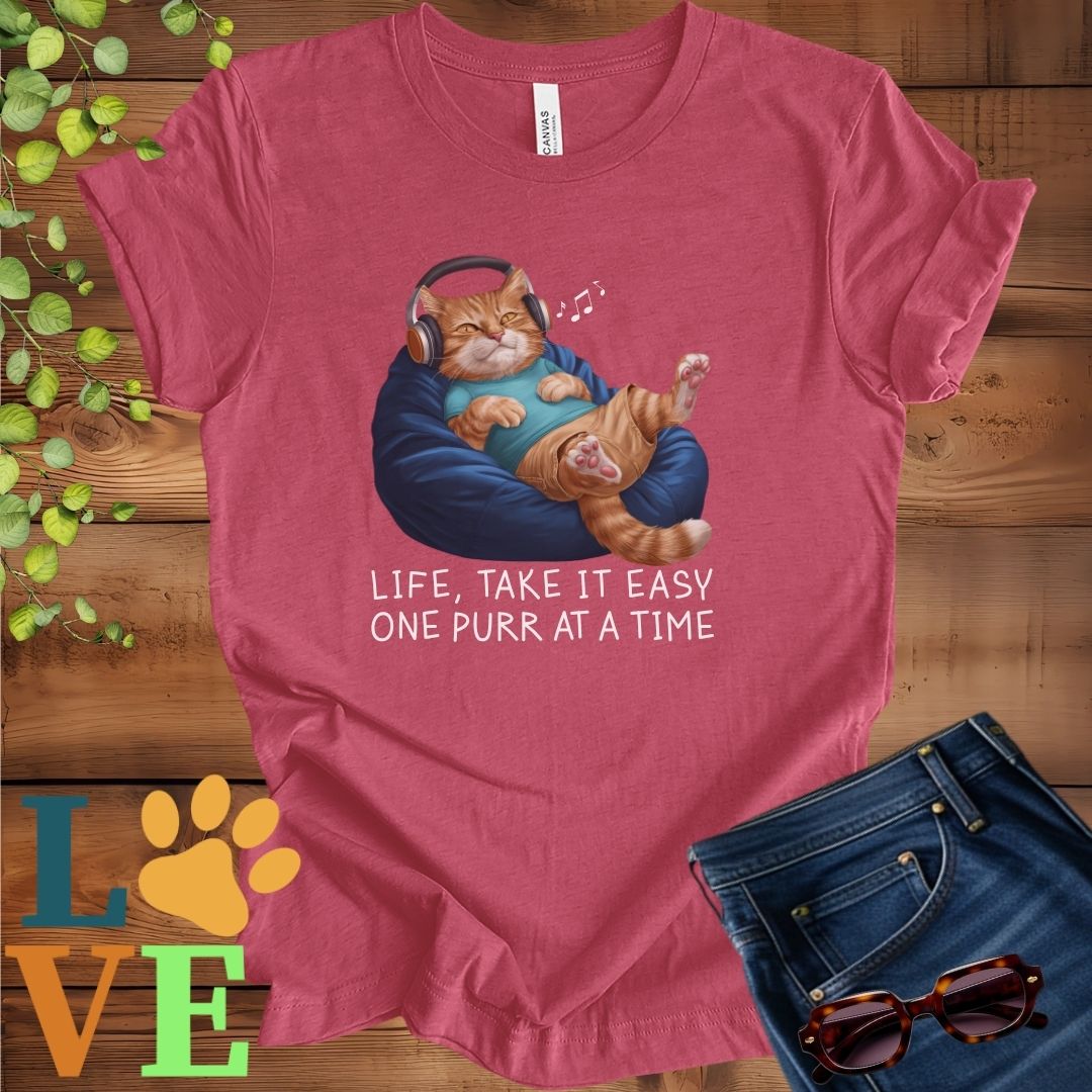Muted raspberry pink (heather raspberry) t-shirt featuring an anthropomorphic ginger cat relaxing on a bean bag while listening to music through headphones, with the slogan "Life, take it easy. One purr at a time."
