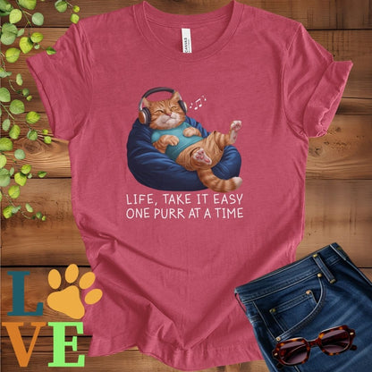 Muted raspberry pink (heather raspberry) t-shirt featuring an anthropomorphic ginger cat relaxing on a bean bag while listening to music through headphones, with the slogan "Life, take it easy. One purr at a time."
