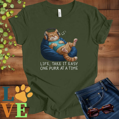 Olive green (military green) t-shirt featuring an anthropomorphic ginger cat relaxing on a bean bag while listening to music through headphones, with the slogan "Life, take it easy. One purr at a time."