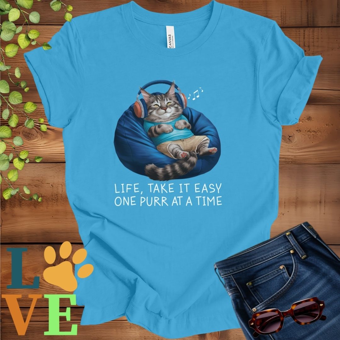 Bright aqua blue t-shirt featuring an anthropomorphic Maine Coon cat relaxing on a bean bag while listening to music through headphones, with the slogan "Life, take it easy. One purr at a time."