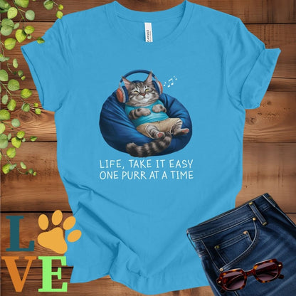 Bright aqua blue t-shirt featuring an anthropomorphic Maine Coon cat relaxing on a bean bag while listening to music through headphones, with the slogan "Life, take it easy. One purr at a time."