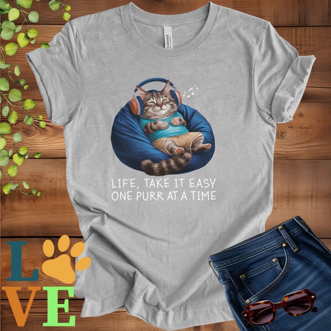 Light grey heather (athletic heather) t-shirt featuring an anthropomorphic Maine Coon cat relaxing on a bean bag while listening to music through headphones, with the slogan "Life, take it easy. One purr at a time."