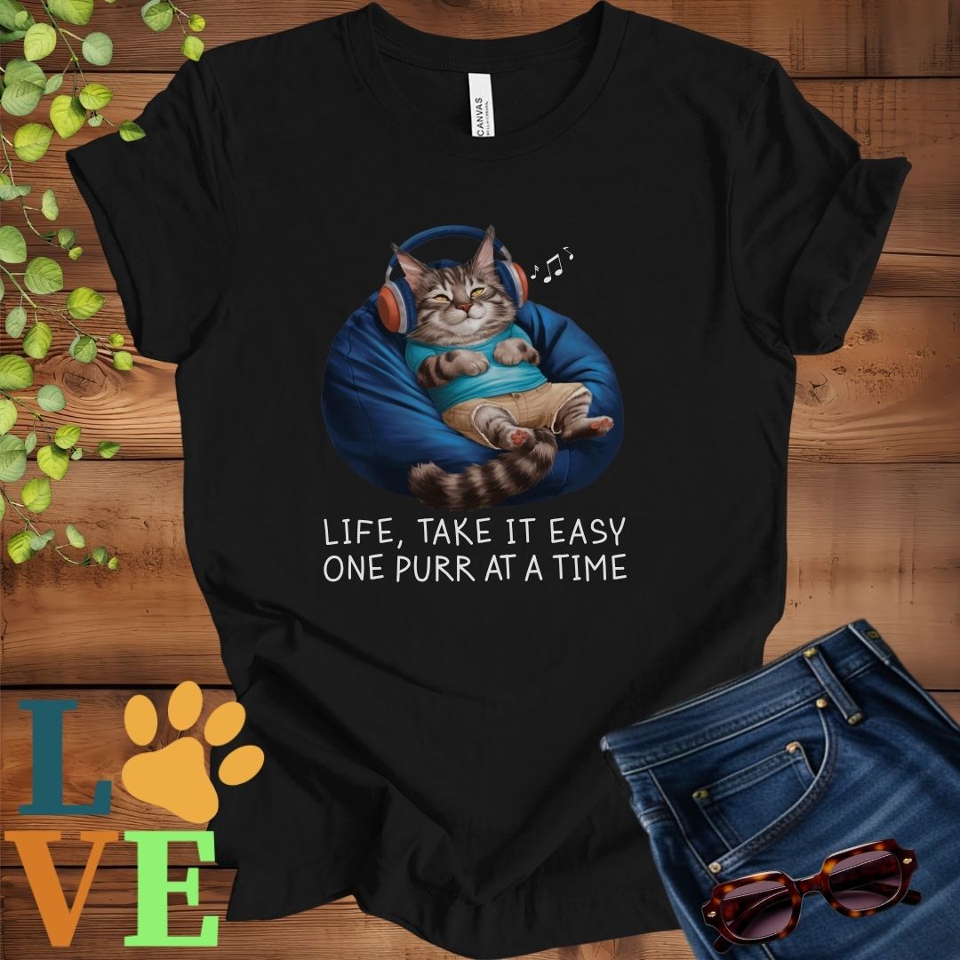 Black t-shirt featuring an anthropomorphic Maine Coon cat relaxing on a bean bag while listening to music through headphones, with the slogan "Life, take it easy. One purr at a time."