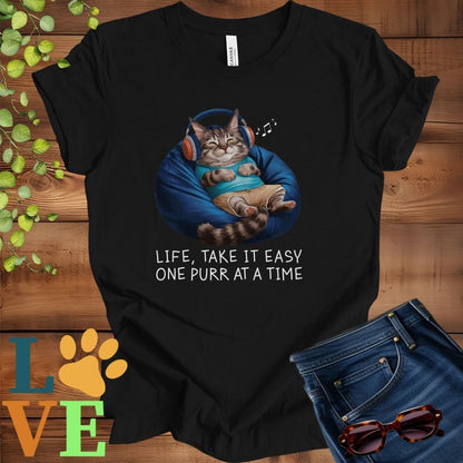 Black t-shirt featuring an anthropomorphic Maine Coon cat relaxing on a bean bag while listening to music through headphones, with the slogan "Life, take it easy. One purr at a time."