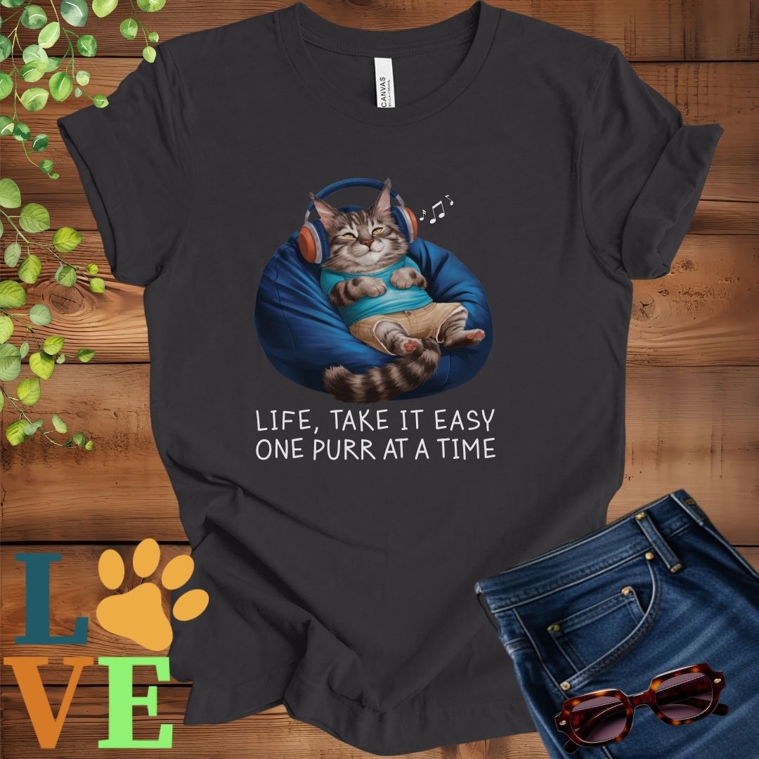 Dark grey heather t-shirt featuring an anthropomorphic Maine Coon cat relaxing on a bean bag while listening to music through headphones, with the slogan "Life, take it easy. One purr at a time."
