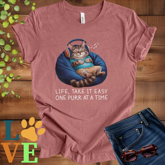 Muted mauve-purple (heather mauve) t-shirt featuring an anthropomorphic Maine Coon cat relaxing on a bean bag while listening to music through headphones, with the slogan "Life, take it easy. One purr at a time."