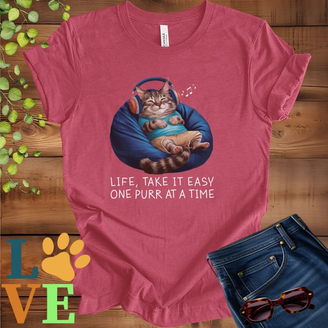 Muted raspberry pink (heather raspberry) t-shirt featuring an anthropomorphic Maine Coon cat relaxing on a bean bag while listening to music through headphones, with the slogan "Life, take it easy. One purr at a time."