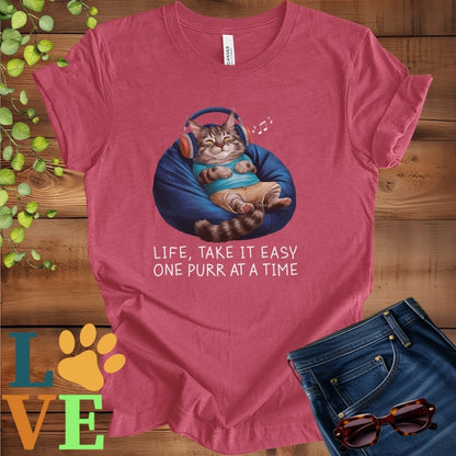 Muted raspberry pink (heather raspberry) t-shirt featuring an anthropomorphic Maine Coon cat relaxing on a bean bag while listening to music through headphones, with the slogan "Life, take it easy. One purr at a time."