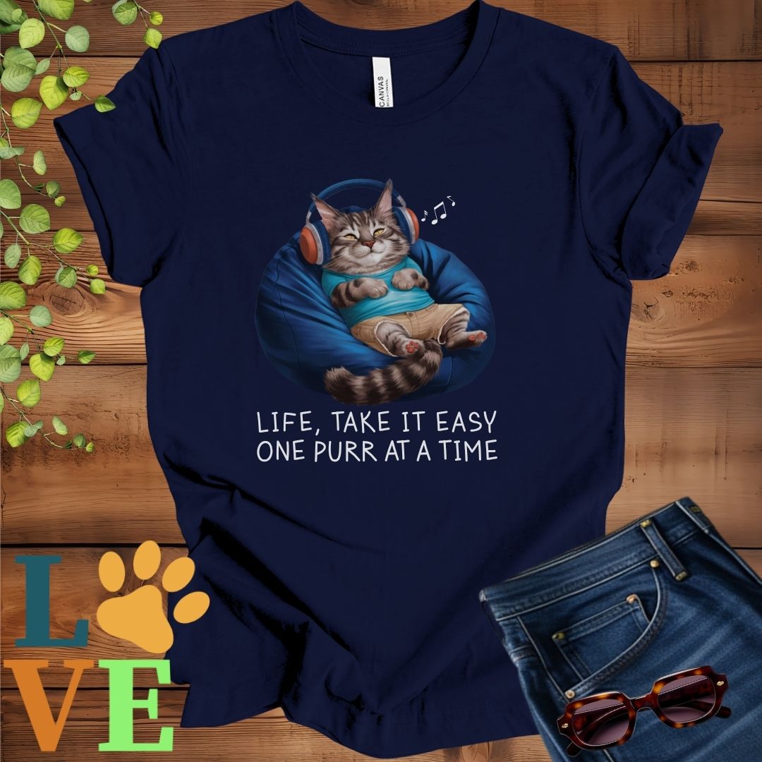 Navy blue t-shirt featuring an anthropomorphic Maine Coon cat relaxing on a bean bag while listening to music through headphones, with the slogan "Life, take it easy. One purr at a time."