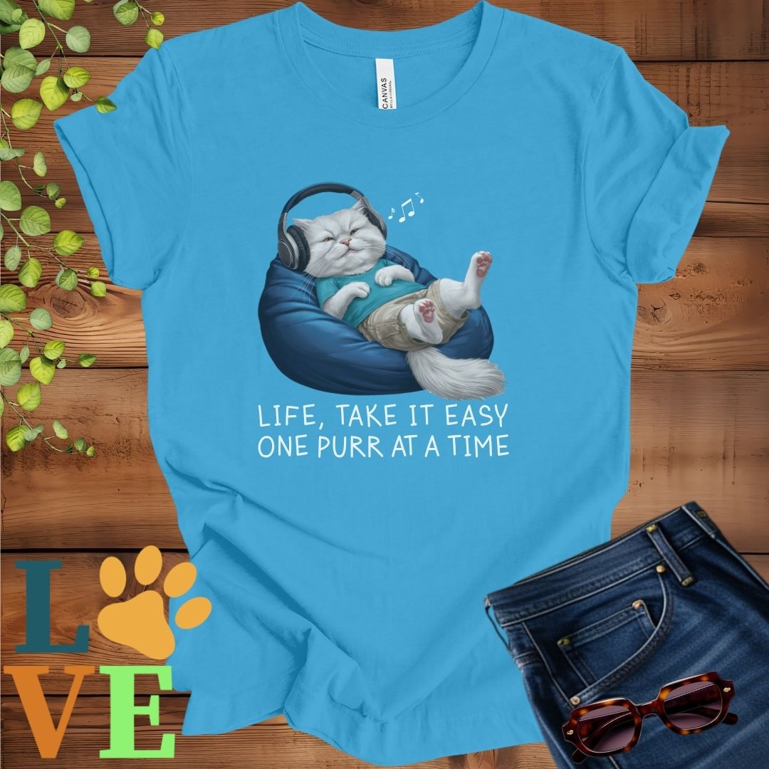 Bright aqua blue t-shirt featuring an anthropomorphic Persian cat relaxing on a bean bag while listening to music through headphones, with the slogan "Life, take it easy. One purr at a time."
