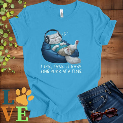 Bright aqua blue t-shirt featuring an anthropomorphic Persian cat relaxing on a bean bag while listening to music through headphones, with the slogan "Life, take it easy. One purr at a time."
