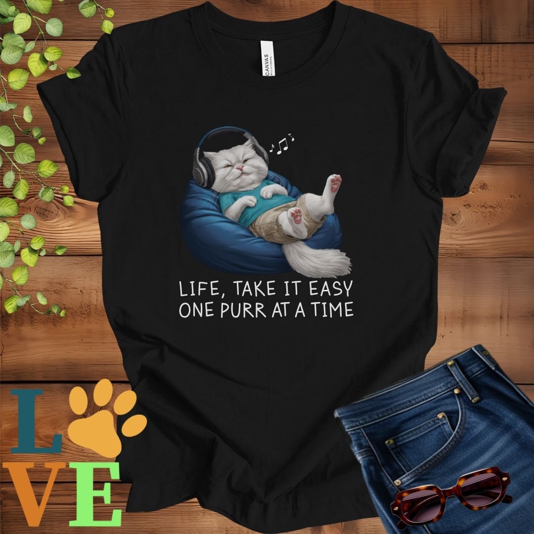 Black t-shirt featuring an anthropomorphic Persian cat relaxing on a bean bag while listening to music through headphones, with the slogan "Life, take it easy. One purr at a time."
