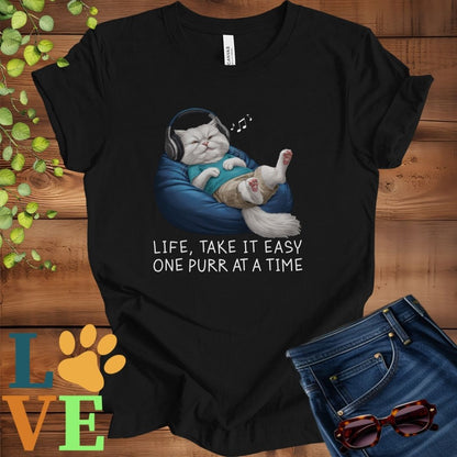 Black t-shirt featuring an anthropomorphic Persian cat relaxing on a bean bag while listening to music through headphones, with the slogan "Life, take it easy. One purr at a time."
