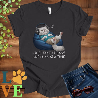 Dark grey heather t-shirt featuring an anthropomorphic Persian cat relaxing on a bean bag while listening to music through headphones, with the slogan "Life, take it easy. One purr at a time."