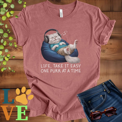 Muted mauve-purple (heather mauve) t-shirt featuring an anthropomorphic Persian cat relaxing on a bean bag while listening to music through headphones, with the slogan "Life, take it easy. One purr at a time."