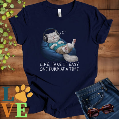 Navy blue t-shirt featuring an anthropomorphic Persian cat relaxing on a bean bag while listening to music through headphones, with the slogan "Life, take it easy. One purr at a time."
