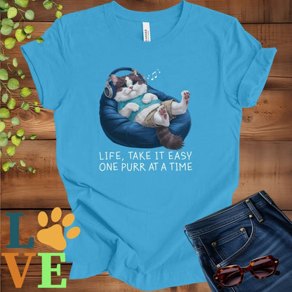Bright aqua blue t-shirt featuring an anthropomorphic Ragdoll cat relaxing on a bean bag while listening to music through headphones, with the slogan "Life, take it easy. One purr at a time."
